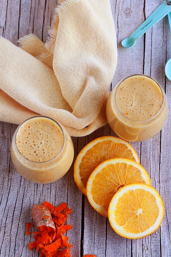 two glasses of orange Julius sitting on top of a wooden table surrounded by slices of fresh oranges