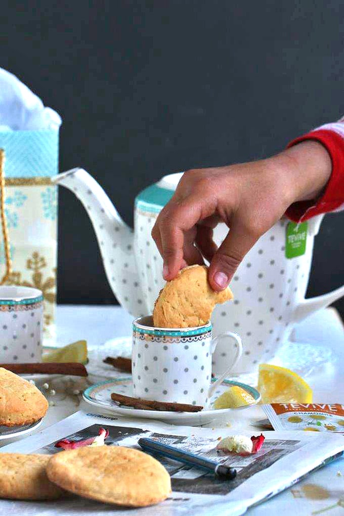 A person dunking a biscuit into cup of tea with tea pot in background