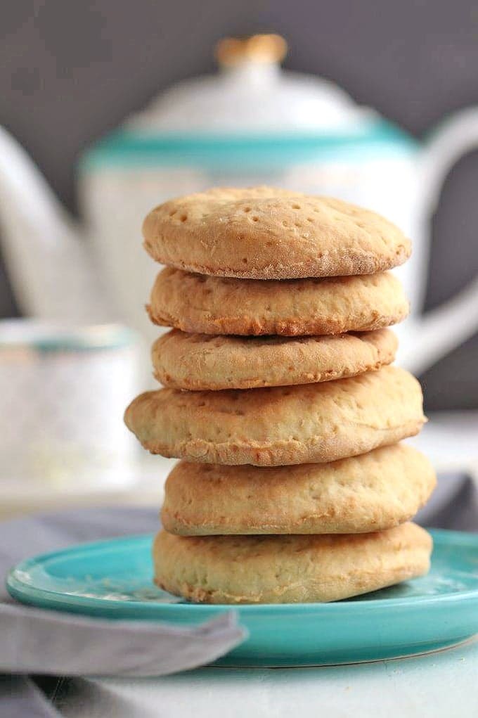 A stack of tea biscuits pancakes on a light blue plate with tea pot in background