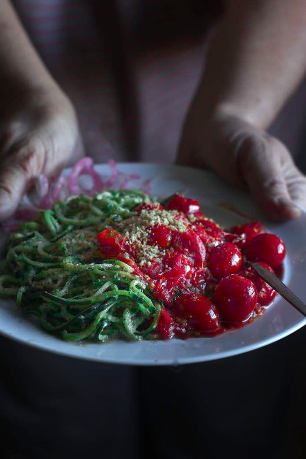 hands holding a plate of pesto zoodles topped with tomatoes