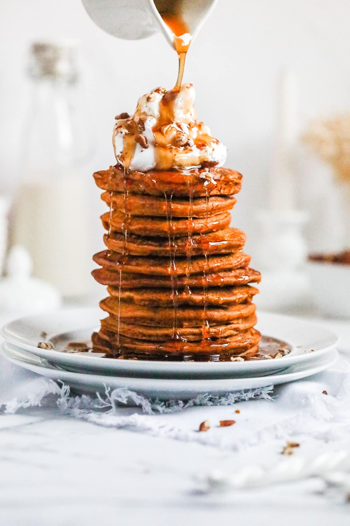 someone pouring maple syrup over a stack of vegan pumpkin pancakes on a white plate