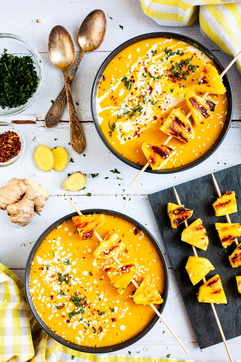 2 bowls of bright yellow soup topped with grilled pineapple skewers